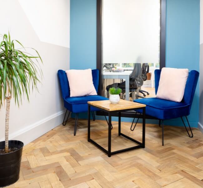 a coworking breakout space including two blue chairs and a table with a plant in the corner